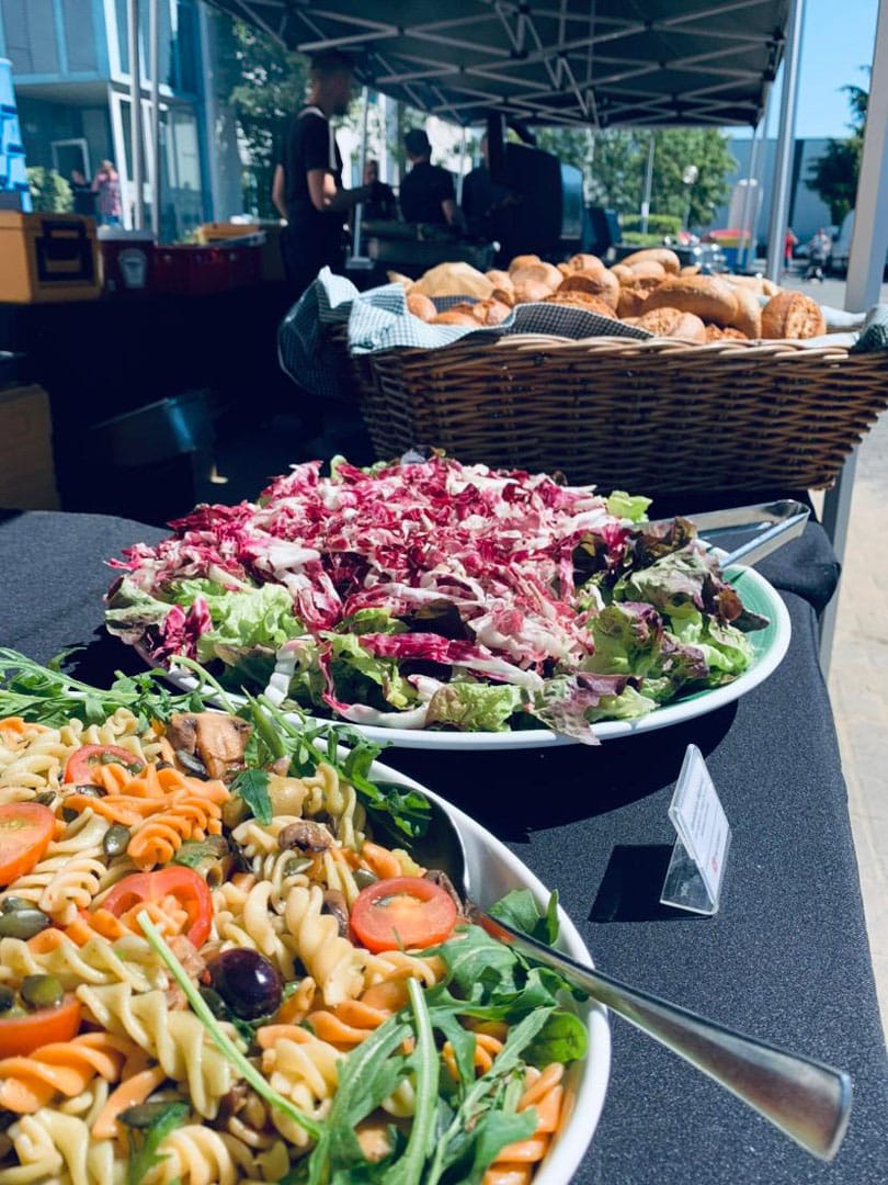 An einer Essensbar steht ein Korb mit Brötchen und zwei Schalen mit einem gemischten Salat und einem Nudelsalat