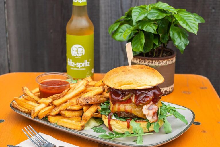 Auf einem Holztisch steht ein Teller mit einem Burger, Pommes Frites mit Sauce im Schälchen; davor liegen auf einer weißen Serviette Messer und Gabel; dahinter steht eine grüne Pflanze und ein Erfrischungsgetränk