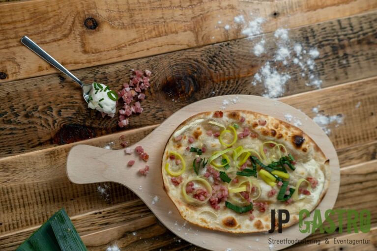 Die Ansicht von oben eines klassichen Flammkuchens Elsässer Art, welcher auf einem Brettchen auf einem Tisch steht; umgeben von einem Löffel mit Schmand, Frühlingszwiebeln und Speck