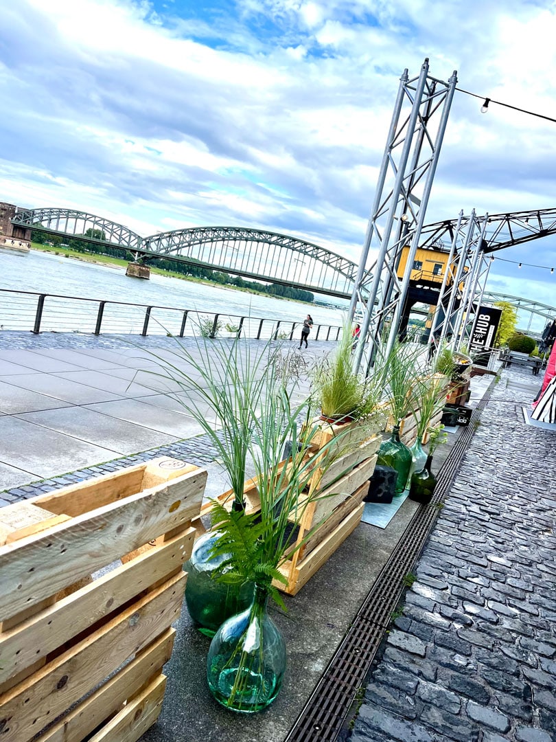 Mehrere dekorative Elemente aus Holzpaletten & gläsernen Bauchflaschen mit Pflanzen in einer Reihe an einem großen Fluss mit einer Bogenbrücke im Hintergrund