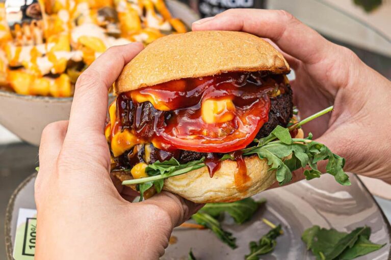 Ein Gast, der den veganen Burgern mit beiden Händen festhält; im Hintergrund weiteres Essen und Getränkeflaschen